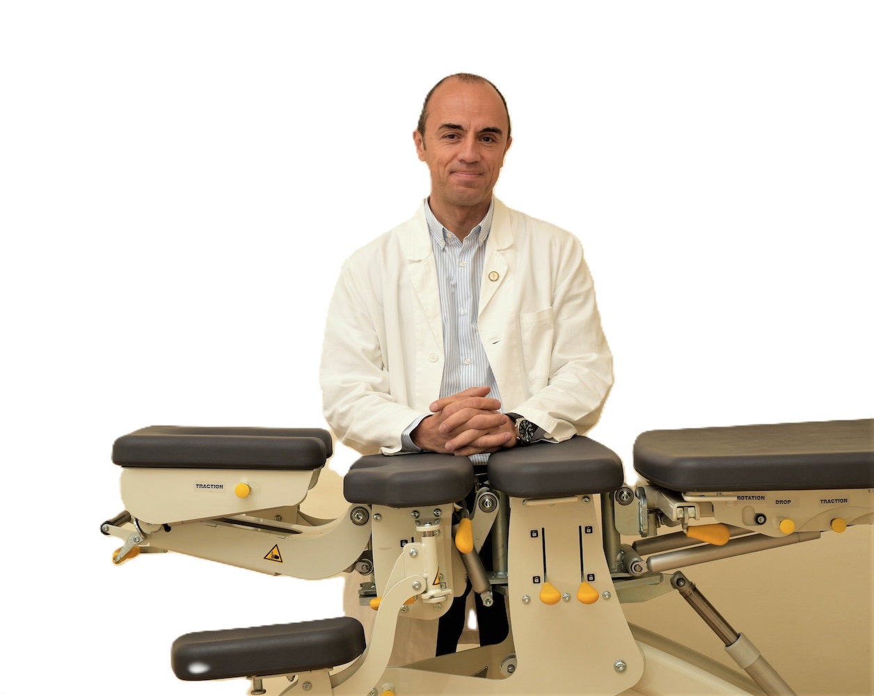 Nell’immagine il Dott. Giancarlo Viano seduto dietro un lettino chiropratico professionale. Indossa un camice bianco e una camicia chiara, con un’espressione serena e professionale. Le mani sono poggiate con calma sul lettino, in una postura accogliente e rassicurante.<br />
Il lettino presenta diverse sezioni regolabili e comandi visibili, tipici degli strumenti utilizzati in chiropratica e trattamenti manuali.